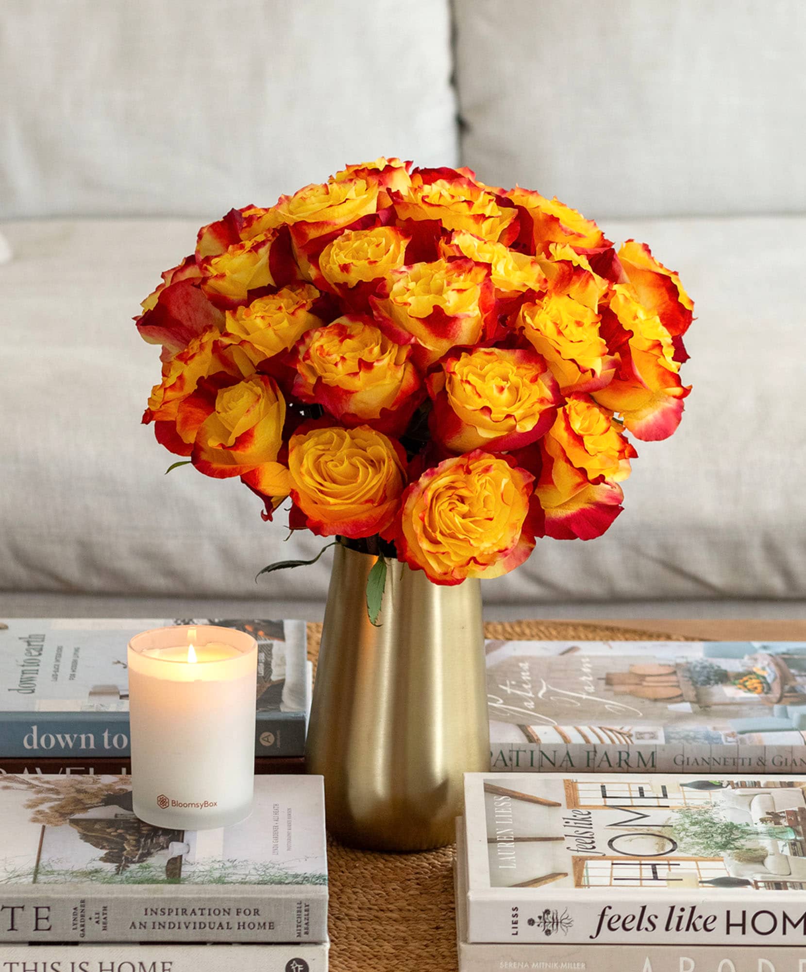 Vibrant orange and yellow rose bouquet in a stylish gold vase, surrounded by books and a candle.